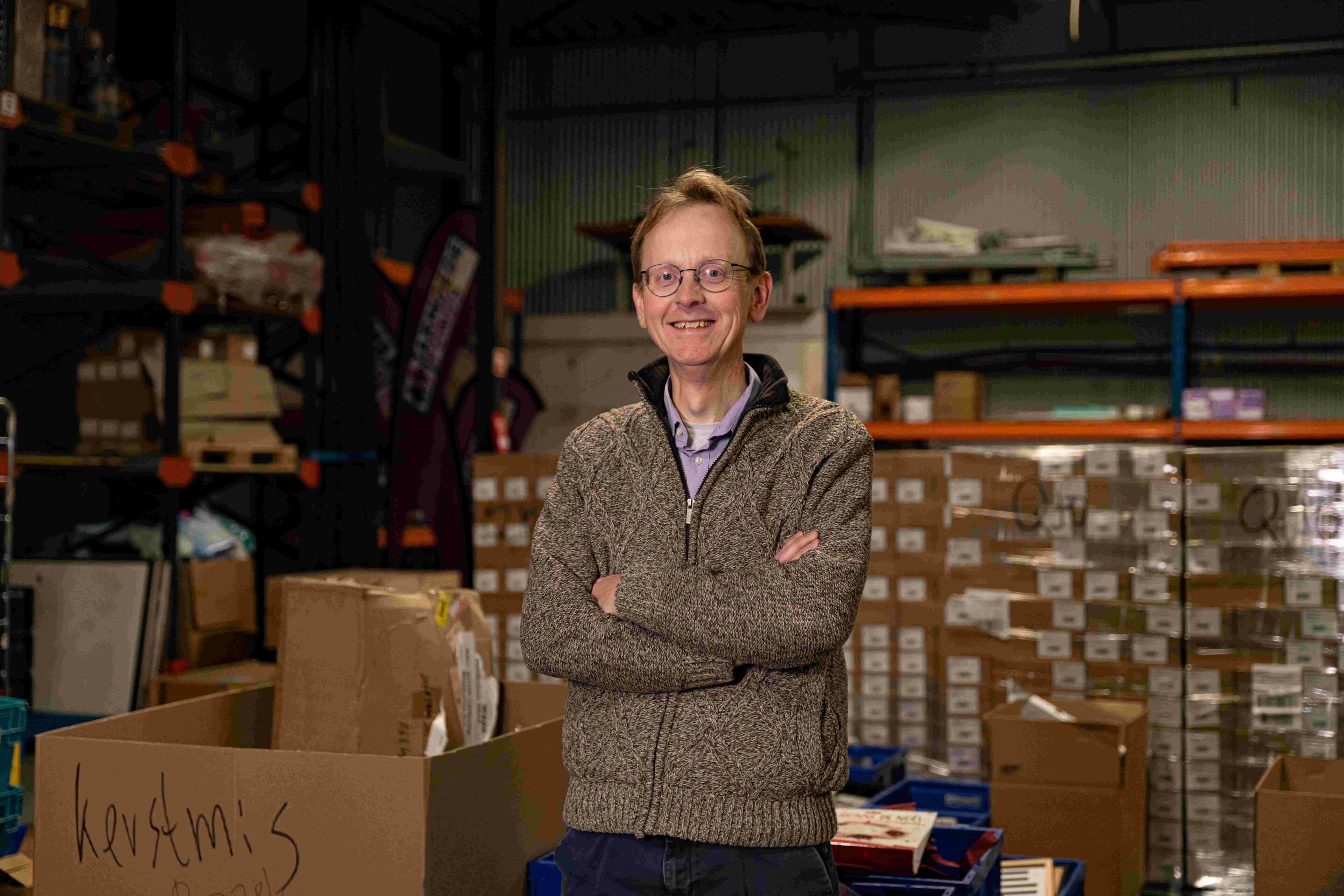 Henk de Graaf profiel website Henk de Graaf in de loods van het Armoedefonds. Er staan veel pallets met goederen op de achtergrond.